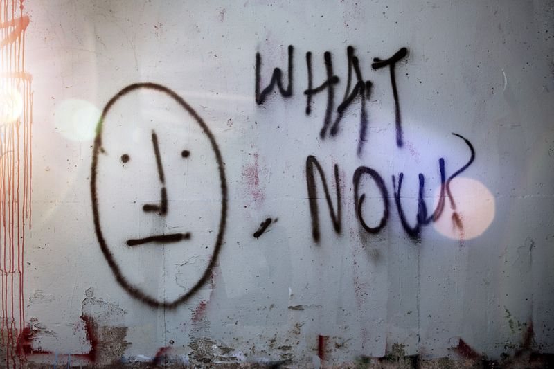 wall with peeling paint covered by a large spray painted face with a neutral expression, graffiti text reads 'what now?'
