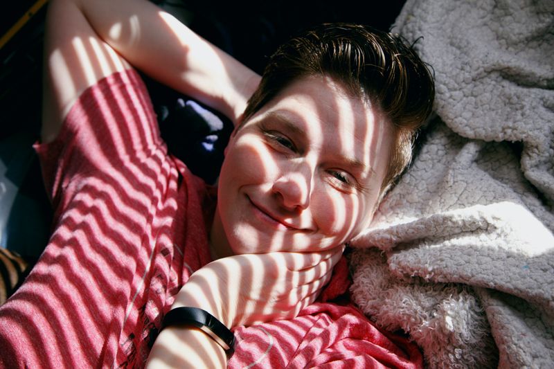 A person lying down on a bed, with a warm smile under sunlight coming through the blinds.