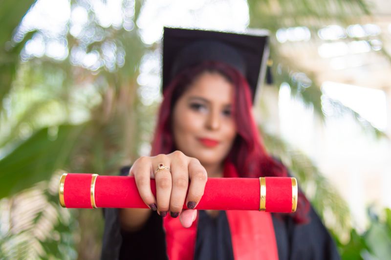 Person with long red hair, a graduation cap and a gold ring holds a red and gold diploma in the foreground