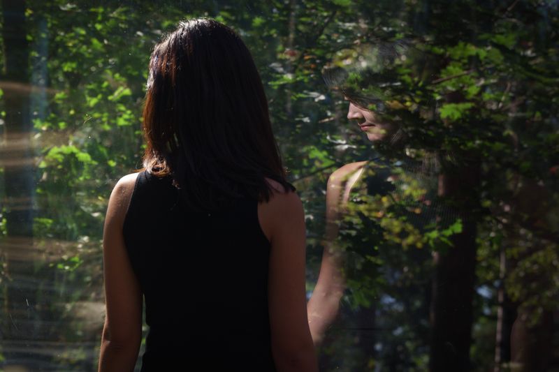 A person looks out a window at a forest. Their reflection is visible in the glass.