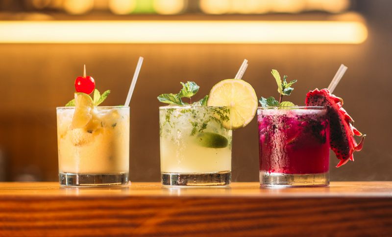 Three cocktails: yellow with a cherry, lime with mint, and pink with dragon fruit, on a wooden surface.