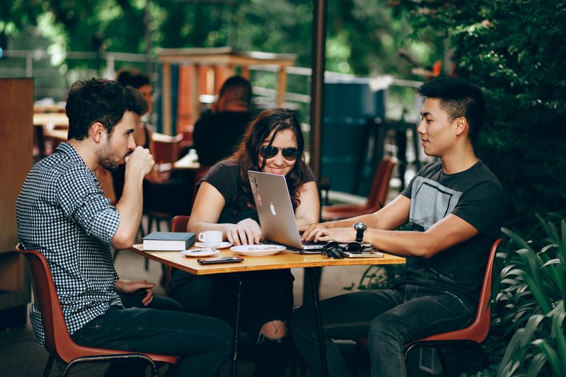 Photo of a three young people, two males and a female, sitting around a table outdoors drinking coffee & working on a laptop