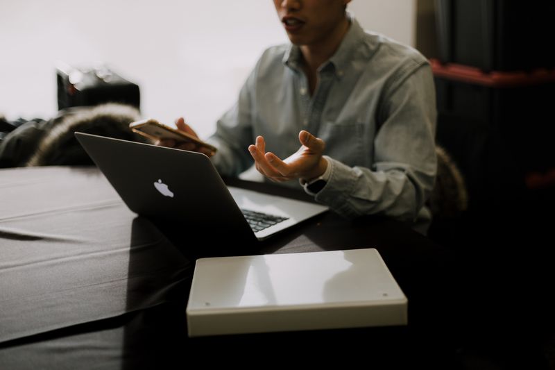 Man appears to be explaining something to someone while on his phone and computer.