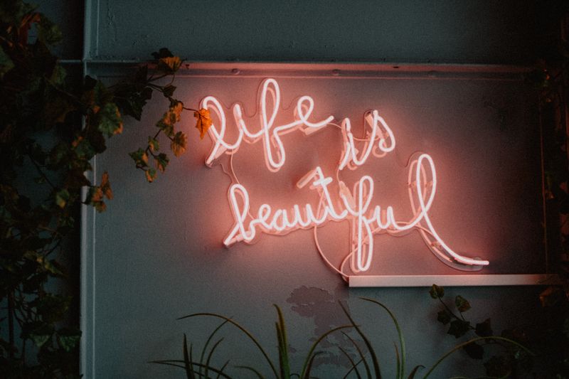 Pink neon sign that says, 'life is beautiful.' While surrounded by plants and hanging on a wall.