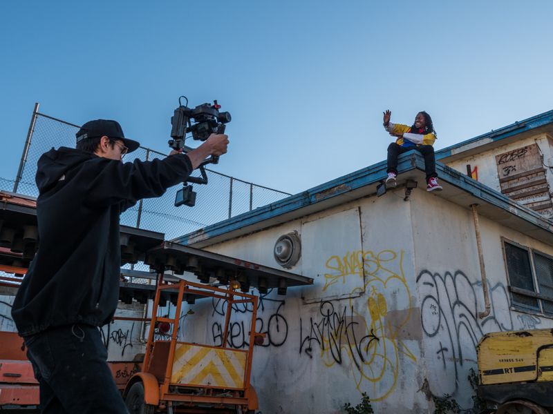 Image with a cameraman pointing a camera at a woman sitting on the roof of a building.