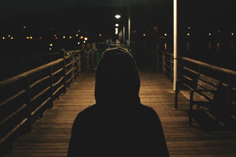 A person wearing a hoodie walks down a boardwalk at night.