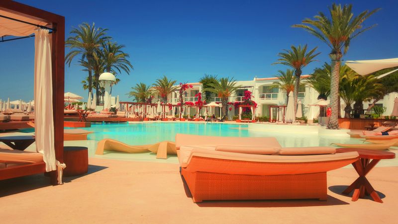 A luxurious hotel with a pool surrounded by lush palm trees and sun lounger chairs.