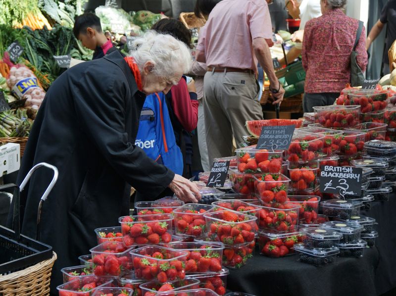 An old lady in a supermarket is choosing some berries. There are punnets of strawberries, blackberries and blueberries.