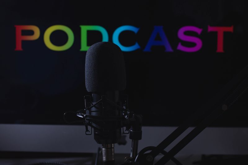 Photo of a microphone and stand against a black background with the words Podcast capitalized in florescent lights