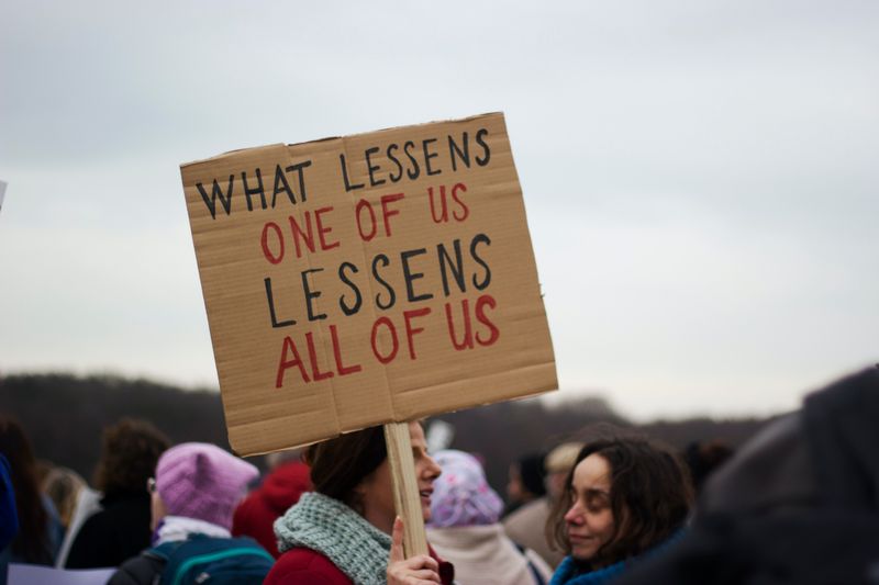 A woman holding a sign that says, 'What Lessens One of Us Lessens All of US.'