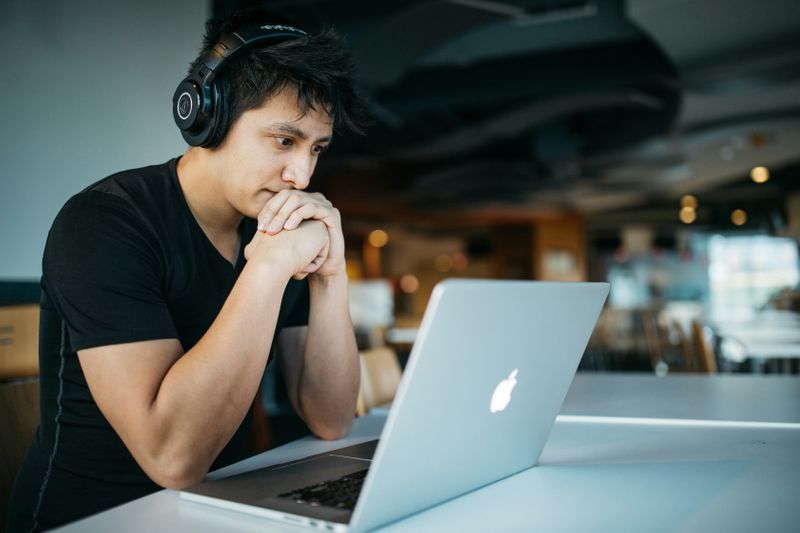 A man listens to headphones as he works on a laptop in a cafe.