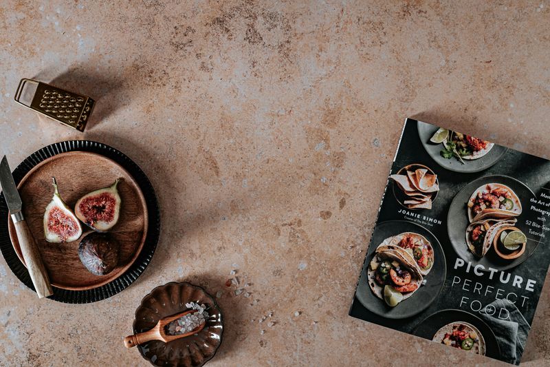 A cookbook and utensils on a table beside figs and salt on wooden plates.