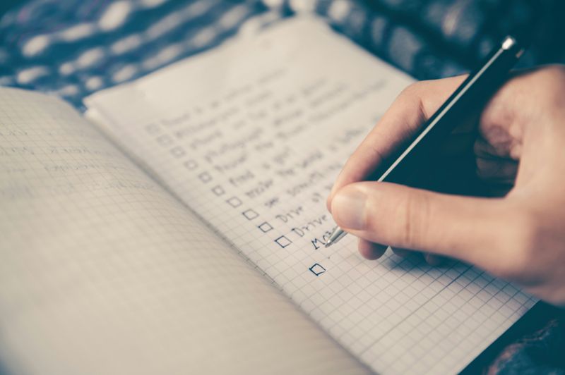 A hand holding a pen, writing on a checklist in a notebook.