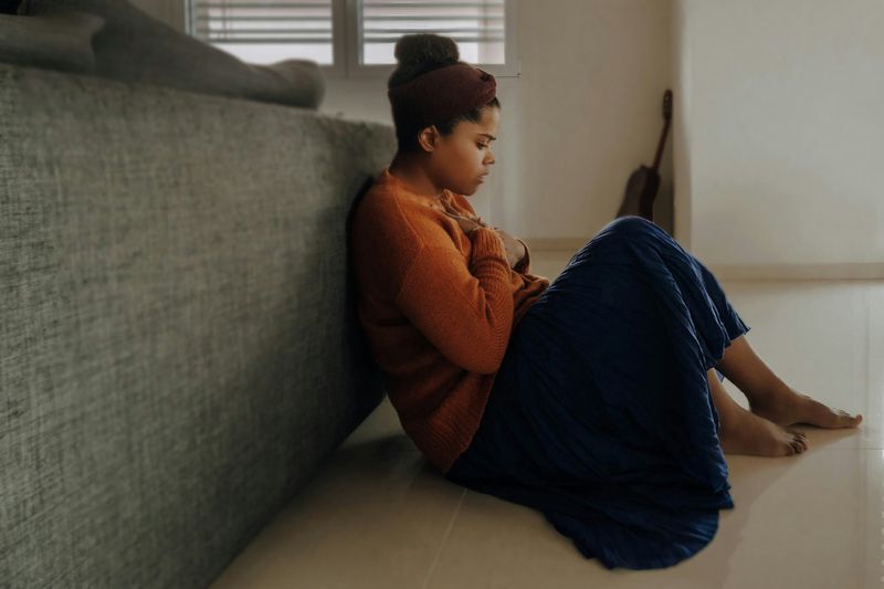 A young woman sitting on the floor with a worried expression, hands on her chest.