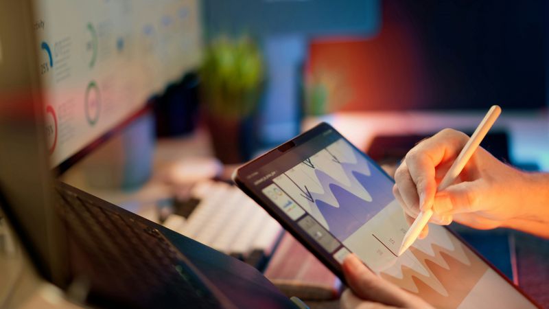 A person on tablet with computer monitor in the background with charts and graphs.