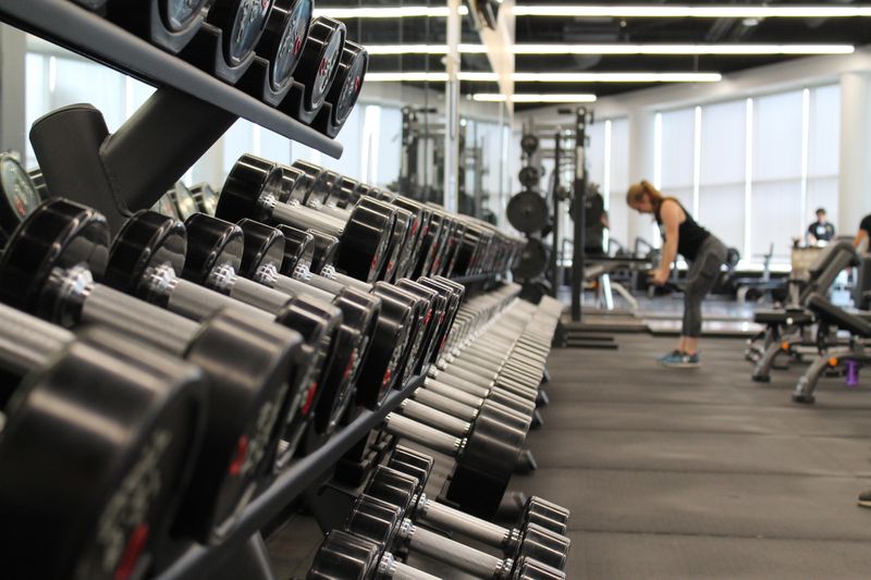 Image of a gym with weight rack in the foreground. A woman is working out in the background.