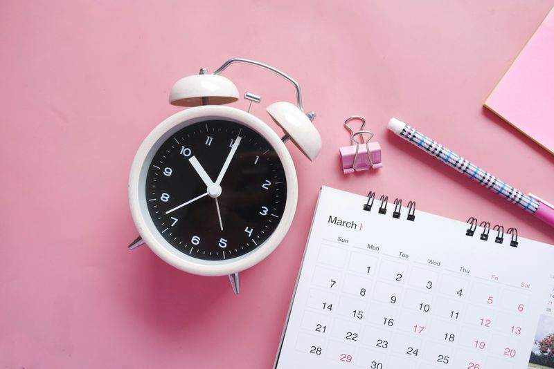 An analog ring clock sitting on a pink background and next to a white paper calendar.