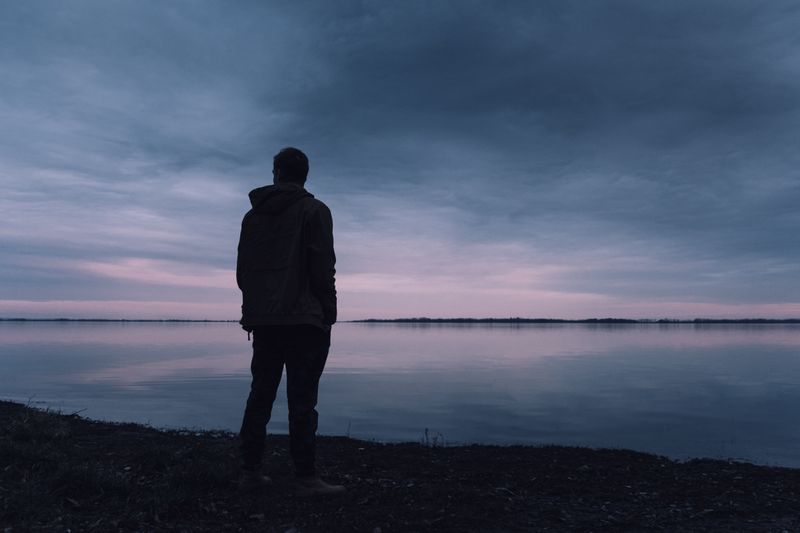 Man standing by the lake looking out with dark clouds above him at dusk.