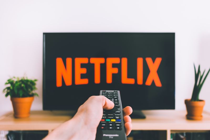 A hand holding remote in the center with Netflix logo on the TV screen. Plants sit on the left and right of the tv.