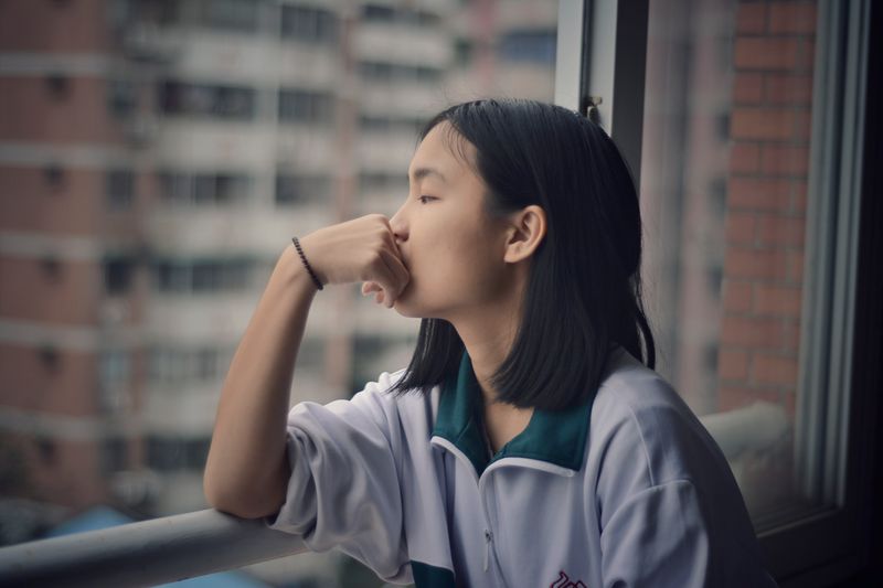Asian woman starts out window in deep thought. She has one hand propped up against her chin. 