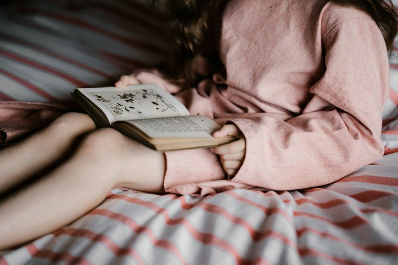 A young girl sitting on a bed reading a book.