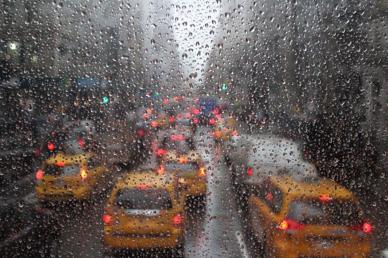 A window of a bus covered in raindrops, overlooking a city street crowded with yellow taxis in traffic.