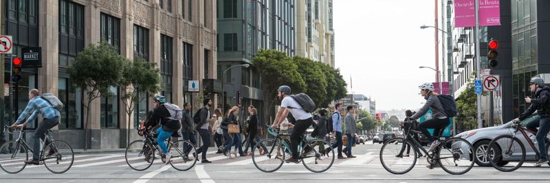 An intersection in a busy city. Cars, bikes, and pedestrians all cross the street.