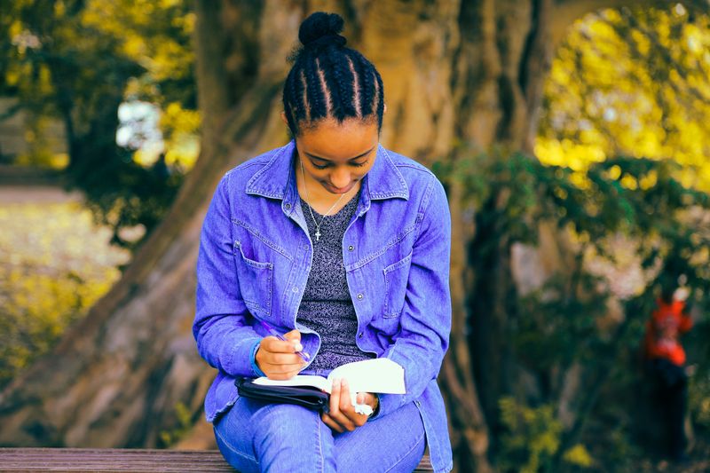 A woman sitting on a bench outdoors writing in a notebook.