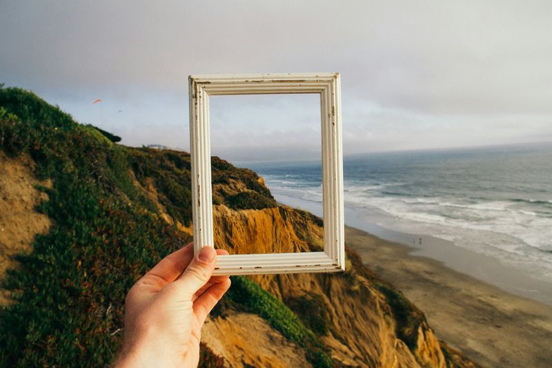 A person holding up an empty frame in front of a scene that includes a rocky hill above a beach. 