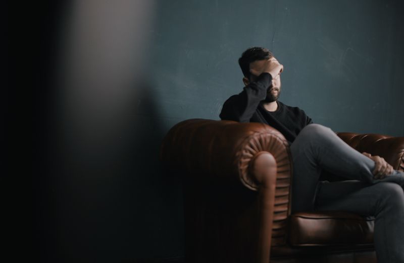 A distressed man is seated on a sofa, pressing his right hand against his temples in a gesture of discomfort or worry.