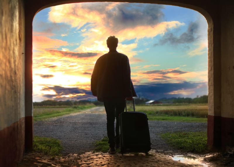 A silhouette of a person with a carryon bag walking forward into a beautiful landscape.