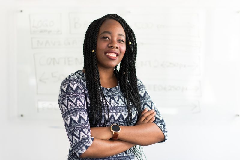 woman who is black with long braids, smiling with arms folded, standing in front of a whiteboard with faint writing