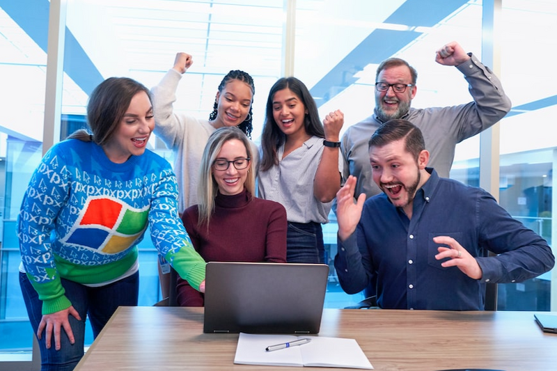 A group of people are looking happily at a laptop screen. Some of them have fists in the air like saying 