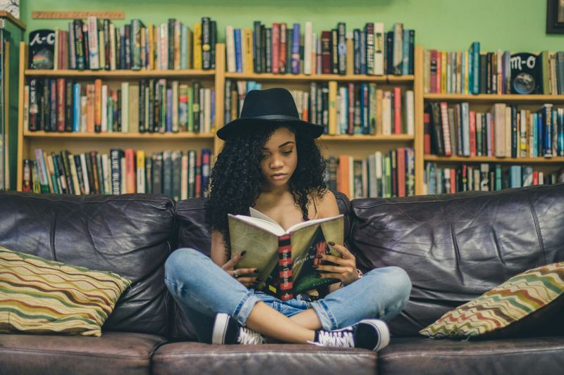 A girl reading books on a couch. Booksheleves filled with books are in the background.