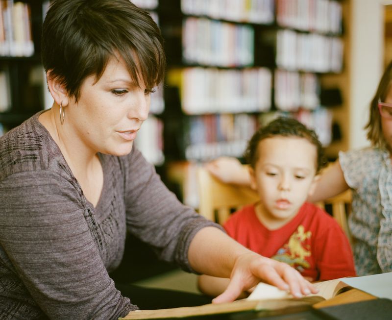 A woman looking at a book and reading it out loud to two young kids.