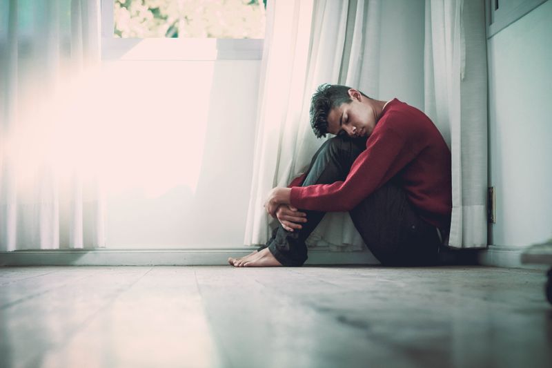 Anxious young man is curled up on the floor in an anxious pose cradling his knees. 