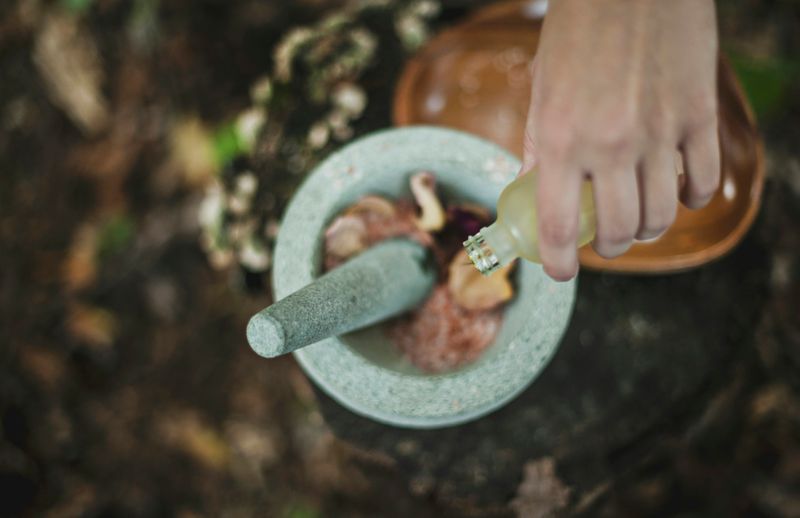 A hand pouring a bottle into a mortar containing items, with a pestle inside the mortar.