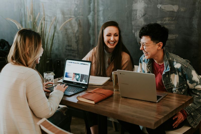 Three young adults sitting around a table with laptops and notebooks open. They smile as they work together.