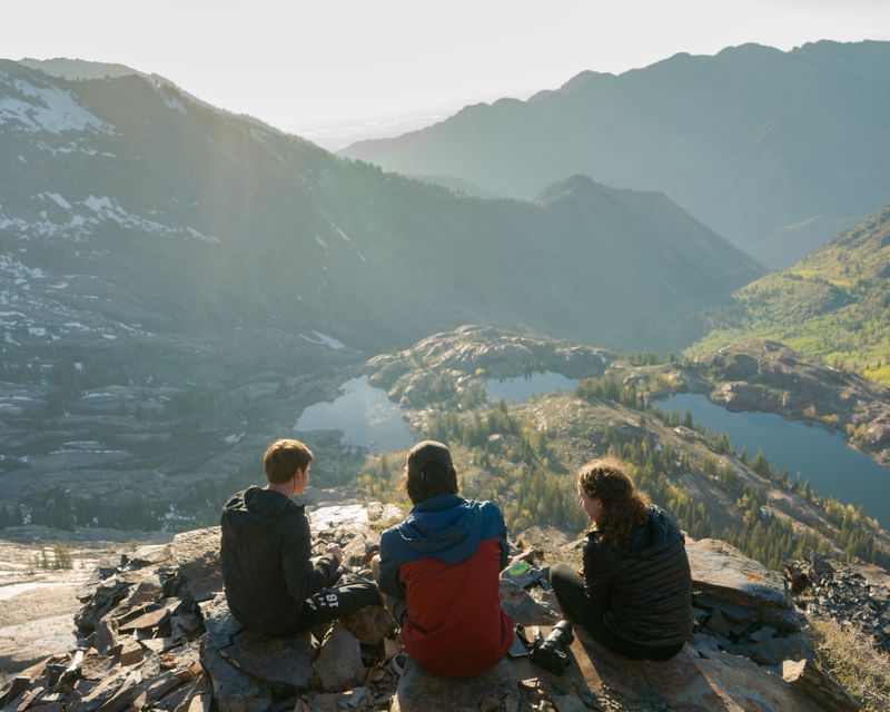 Image of three people (2 male and 1 female) sitting on a high rock that overlooks a valley surrounded by mountains