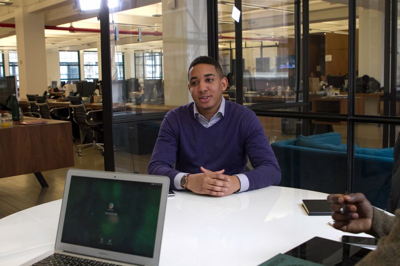 Young man with brown skin and dark hair sitting in an office, opposite an open laptop.