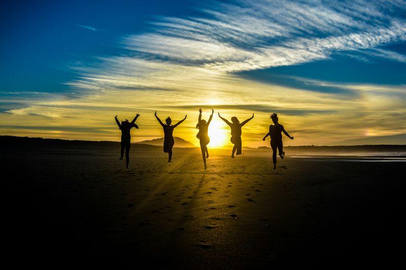 Five people on a beach, jumping with their arms in the air. They're in silhouette with the sun behind them. 