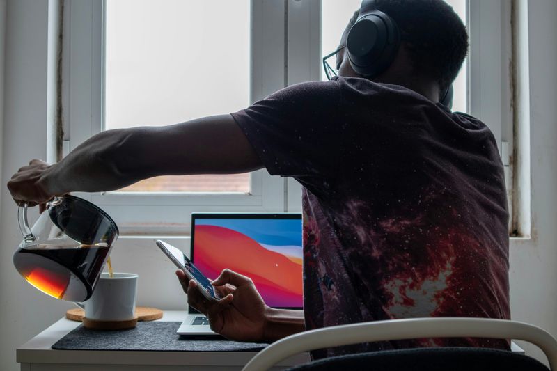 Man sitting in front of his laptop, holding his phone, wearing headphones, and pouring coffee.