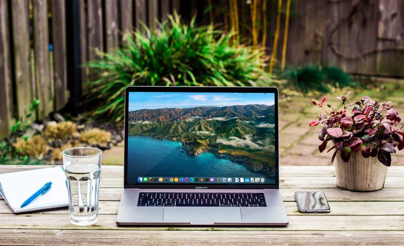 Open laptop computer on outdoor wooden table with pad, blue pen, glass of water, cellphone, & a red-leaf plant in a white pot