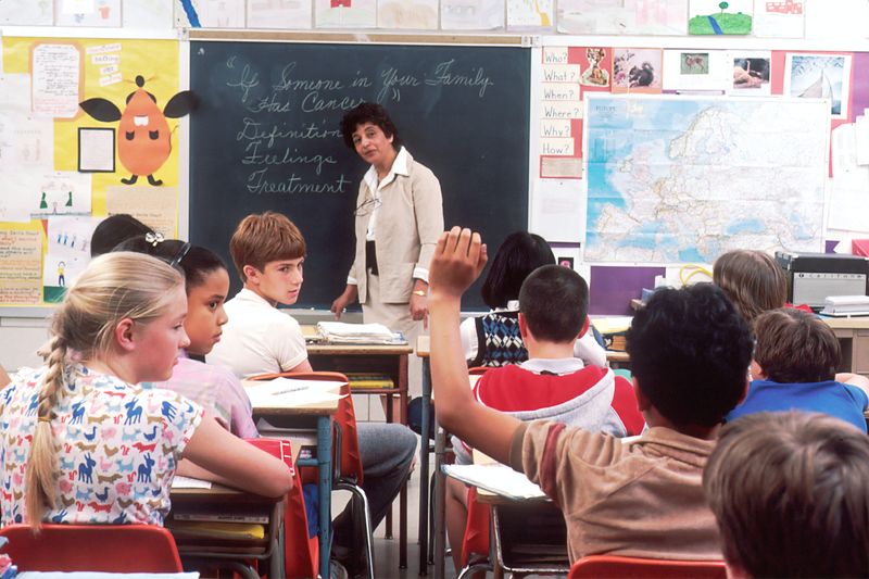 A classroom with a teacher standing by a black board and group of students seated in front of her.