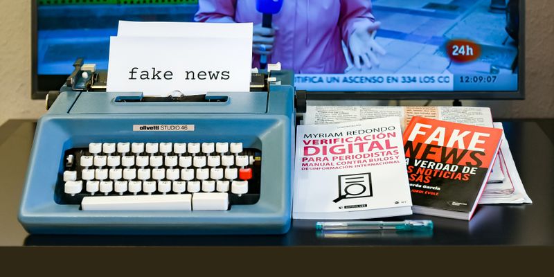 A typewriter and books in front of a screen that have the phrase fake news in them.