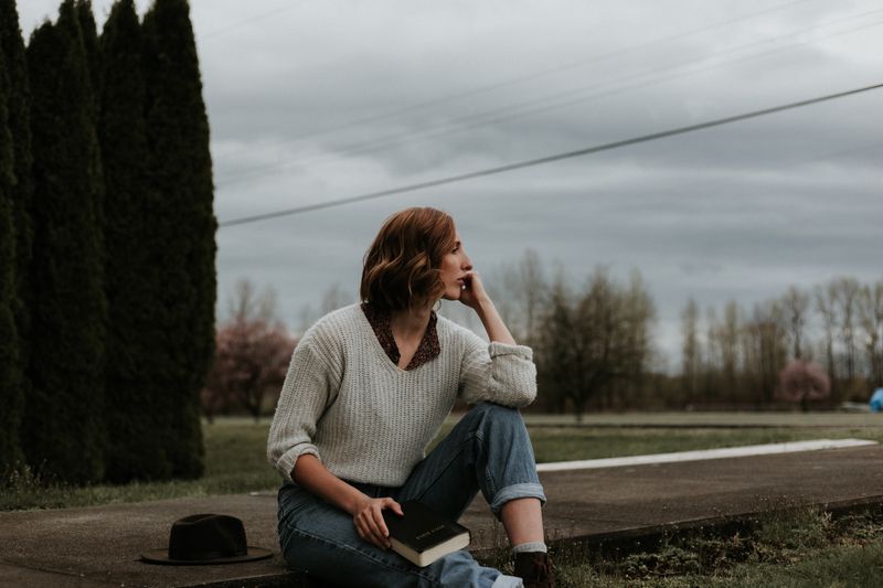 A woman sitting on sidewalk with a book on her leg in deep thought.