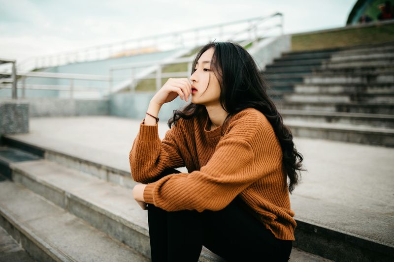 An young woman sits on steps looking out with her hand resting on mouth.