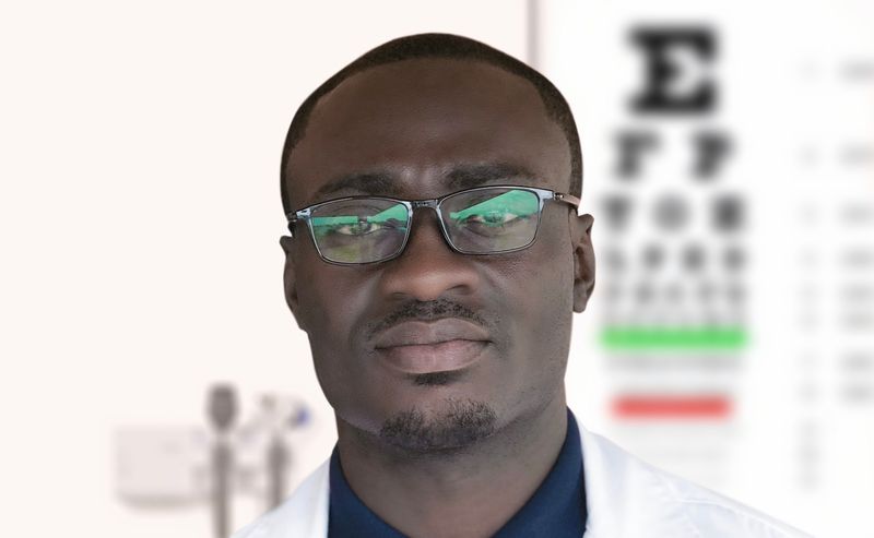 Person, in a white lab coat with glasses on. In the background, there is eye chart on the wall.