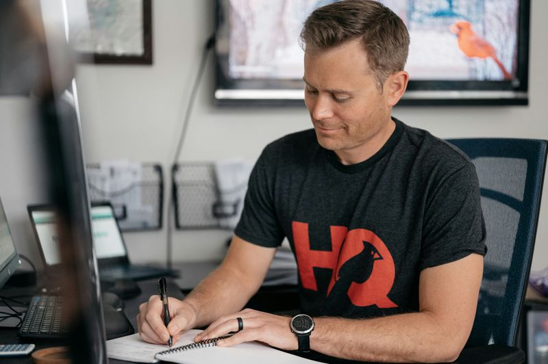 A man sitting at desk in front of a computer writing in a notebook. 