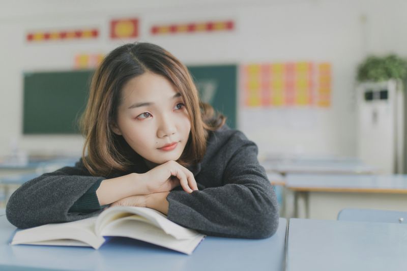 A young woman sitting alone with her hands resting on an open book, and her chin on the back of her hand. 